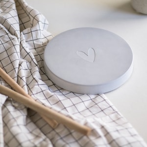 Könnte beinhalten: Ein runder Untersetzer aus hellem Beton mit einem eingegrabenen Herzen. Der Untersetzer liegt auf einem weißen Stoff mit schwarzen Karos.