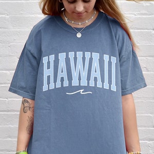 May include: A blue oversized t-shirt with the word "HAWAII" printed in white with a blue wave underneath. The shirt is being worn by a person with long blonde hair.
