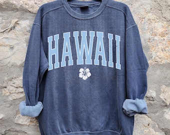 May include: A navy blue sweatshirt with the word "HAWAII" in light blue block letters across the chest. A white hibiscus flower is centered below the text. The sleeves are rolled up, and the sweatshirt hangs on a black hanger.