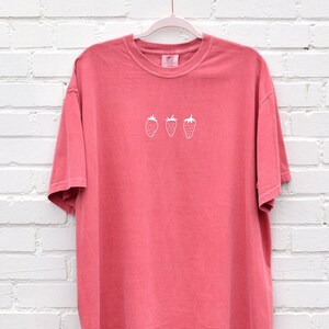 Op de afbeelding: Een koraalroze T-shirt met een wit geborduurd ontwerp van drie aardbeien.