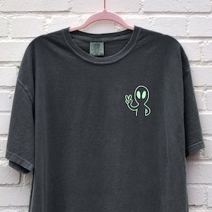 Puede incluir: Una camiseta gris oscuro con un gráfico de extraterrestre verde en la parte delantera. El extraterrestre está haciendo un signo de paz con su mano.