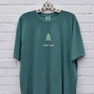 Peut inclure: Un t-shirt vert sarcelle avec un pin de dessin animé et le texte "feelin' pine" en blanc.