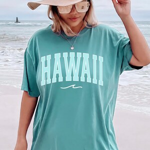 May include: A teal blue oversized t-shirt with the word "HAWAII" printed in a light blue color with a white wave underneath. The shirt is being worn by a person on a beach.