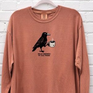 May include: A long-sleeved, burnt orange t-shirt with a black crow illustration holding a cup of coffee. The text "Hello darkness my old friend" is printed below the crow.