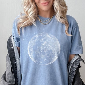 May include: A pale blue t-shirt with a white illustration of the moon. The celestial design is centered on the shirt, with a starry background. The shirt is paired with a dark denim jacket.