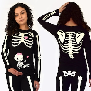 May include: Black long-sleeved shirt with a glow-in-the-dark skeleton design. The shirt features a small skeleton baby in the stomach area with a pink bow and two pink hearts. The back of the shirt shows a full skeleton design.