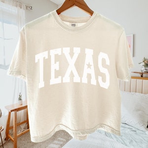 K&ouml;nnte beinhalten: Ein cremefarbenes, bauchfreies T-Shirt mit einem wei&szlig;en "TEXAS"-Aufdruck auf der Vorderseite. Das Shirt hat einen Stern im "A" des Aufdrucks.