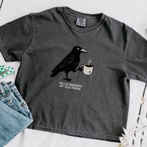 Peut inclure: Un t-shirt gris foncé avec une illustration de corbeau noir tenant une tasse de café. Le texte "HELLO DARKNESS MY OLD FRIEND" est imprimé sous le corbeau.