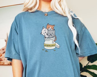 May include: Blue-green t-shirt featuring a graphic of three cartoon cats inside an open prescription bottle labeled "ANTIDEPRESSANTS". The cats are orange, gray, and white. Several yellow pills are scattered around the bottle.