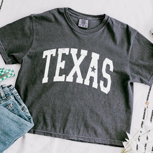 K&ouml;nnte beinhalten: Ein dunkelgraues T-Shirt mit einem wei&szlig;en Vintage-Grafikdruck, auf dem "TEXAS" mit einem Stern &uuml;ber dem "S" steht.