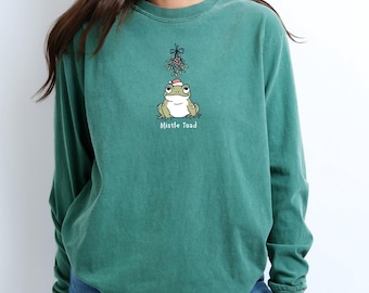 Può includere: Una maglietta verde a maniche lunghe con un disegno di una rana dei cartoni animati che indossa un cappello di Babbo Natale e tiene il vischio. Il testo "Mistle Toad" è stampato sotto la rana.