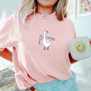 May include: A pink t-shirt with a white goose graphic and the text "Look at all those chickens!" printed on the front. The person wearing the shirt is holding a white mug with a yellow smiley face.