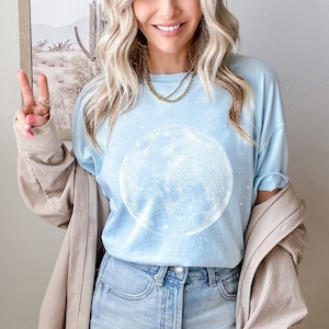 May include: Pale blue t-shirt with a large white moon design. The shirt is worn with blue jeans. A beige jacket is draped over the shoulders. A gold necklace is visible. The image has a casual, relaxed feel.