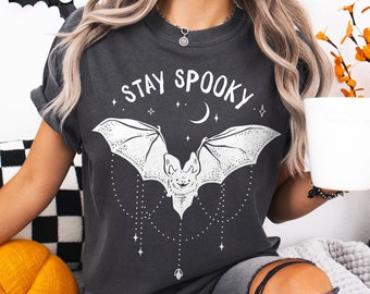 Puede incluir: Una camiseta gris oscuro con un gráfico de murciélago blanco y el texto "Stay Spooky".