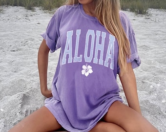 May include: A woman wearing a purple t-shirt with the word "ALOHA" and a white hibiscus flower graphic printed on the front. She is sitting on a sandy beach.
