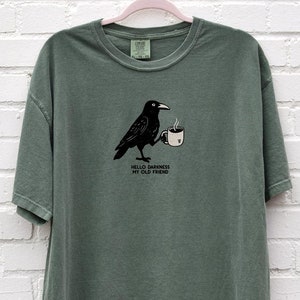 May include: A green t-shirt with a black crow holding a cup of coffee. The text on the shirt reads "Hello darkness my old friend."