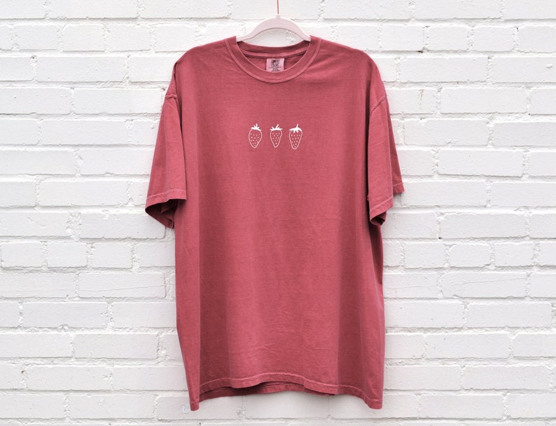 Op de afbeelding: Een rood T-shirt met een wit geborduurd ontwerp van drie aardbeien.