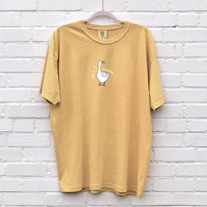 May include: A mustard yellow t-shirt with a white goose graphic and the text "Just a little bit of quacking" printed on the front.