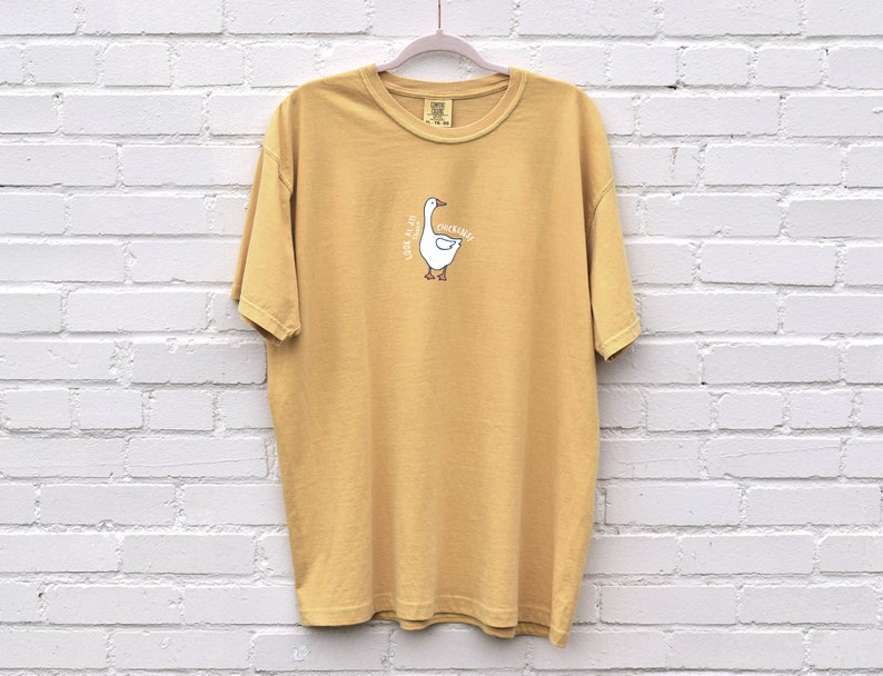 May include: A mustard yellow t-shirt with a white goose graphic and the text "Just a little bit of quacking" printed on the front.