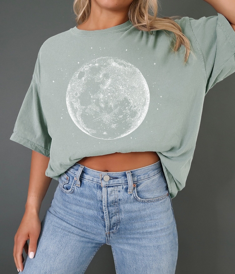 May include: Sage green t-shirt with a white moon illustration and stars. The t-shirt is paired with light blue denim jeans. The moon design is detailed.