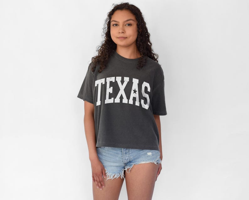 K&ouml;nnte beinhalten: Ein dunkelgraues, bauchfreies T-Shirt mit einem wei&szlig;en Vintage-Style "TEXAS"-Motiv und einem einzelnen wei&szlig;en Stern.