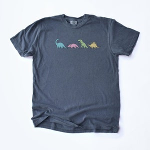 May include: A dark blue t-shirt with a graphic of four dinosaurs in different colors. The dinosaurs are a green brachiosaurus, a pink triceratops, a green tyrannosaurus rex, and a yellow stegosaurus.