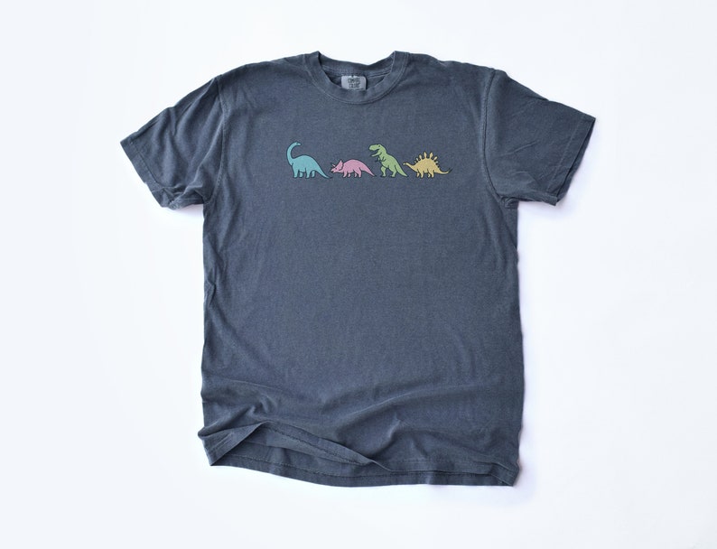 May include: A dark blue t-shirt with a graphic of four dinosaurs in different colors. The dinosaurs are a green brachiosaurus, a pink triceratops, a green tyrannosaurus rex, and a yellow stegosaurus.