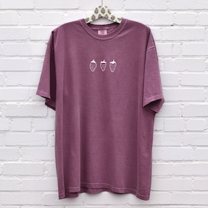 Op de afbeelding: Een mauve t-shirt met een wit geborduurd ontwerp van drie aardbeien.