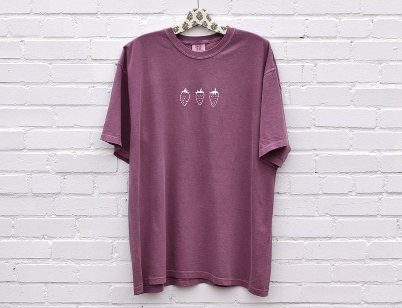 Op de afbeelding: Een mauve t-shirt met een wit geborduurd ontwerp van drie aardbeien.