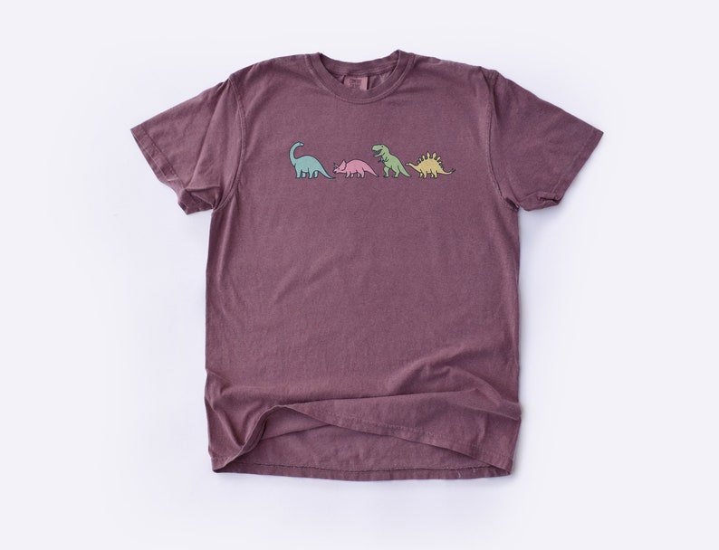 May include: A maroon t-shirt with a graphic of four dinosaurs in a line. The dinosaurs are green, pink, and yellow.