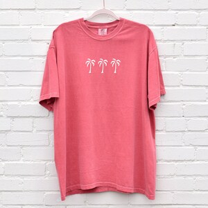 Puede incluir: Una camiseta de color rosa coral con un gr&aacute;fico blanco de tres palmeras. La camiseta tiene cuello redondo y mangas cortas.
