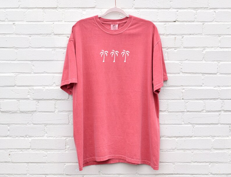 Puede incluir: Una camiseta de color rosa coral con un gr&aacute;fico blanco de tres palmeras. La camiseta tiene cuello redondo y mangas cortas.