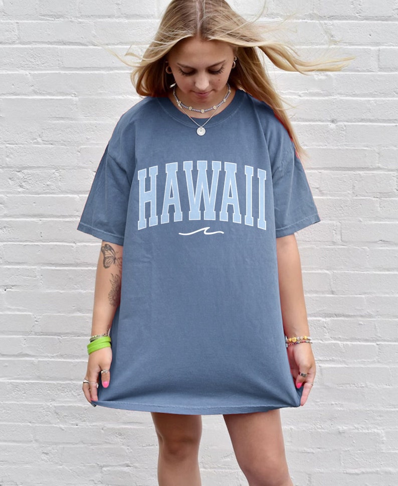May include: A blue oversized t-shirt with the word "HAWAII" printed in white with a blue wave underneath. The shirt is being worn by a person with long blonde hair.