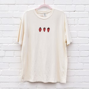 Op de afbeelding: Een cr&egrave;mekleurig T-shirt met een simpel ontwerp van drie rode aardbeien op de voorkant.