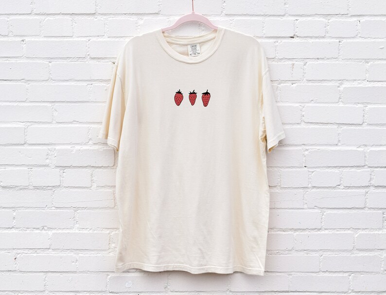 Op de afbeelding: Een cr&egrave;mekleurig T-shirt met een simpel ontwerp van drie rode aardbeien op de voorkant.
