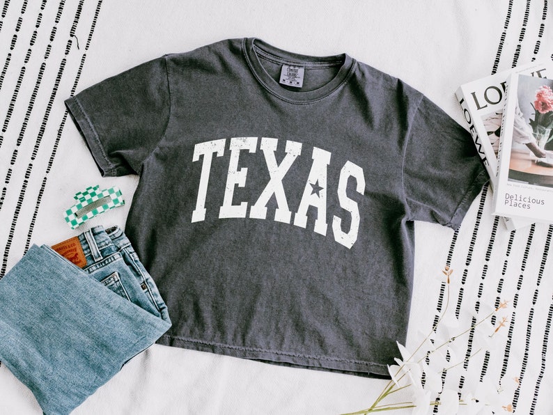 K&ouml;nnte beinhalten: Ein dunkelgraues T-Shirt mit einem wei&szlig;en Vintage-Grafikdruck, auf dem "TEXAS" mit einem Stern &uuml;ber dem "S" steht.
