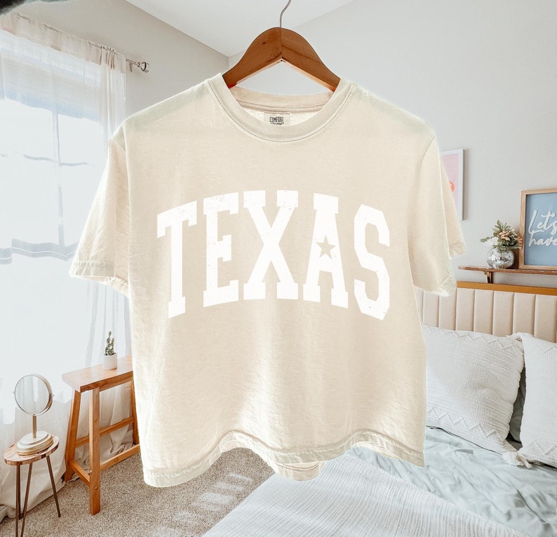 K&ouml;nnte beinhalten: Ein cremefarbenes, bauchfreies T-Shirt mit einem wei&szlig;en "TEXAS"-Aufdruck auf der Vorderseite. Das Shirt hat einen Stern im "A" des Aufdrucks.