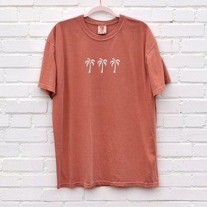 Puede incluir: Una camiseta de color terracota con un gr&aacute;fico blanco de cuatro palmeras. La camiseta tiene cuello redondo y mangas cortas.