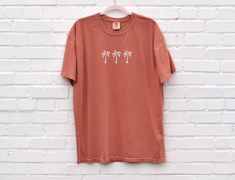 Puede incluir: Una camiseta de color terracota con un gr&aacute;fico blanco de cuatro palmeras. La camiseta tiene cuello redondo y mangas cortas.