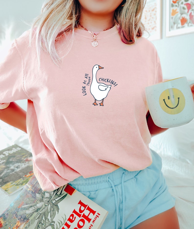 May include: A pink t-shirt with a white goose graphic and the text "Look at all those chickens!" printed on the front. The person wearing the shirt is holding a white mug with a yellow smiley face.