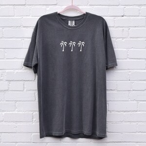 Puede incluir: Una camiseta gris antracita con un gr&aacute;fico de palmera blanca en la parte delantera. La camiseta tiene cuello redondo y mangas cortas.