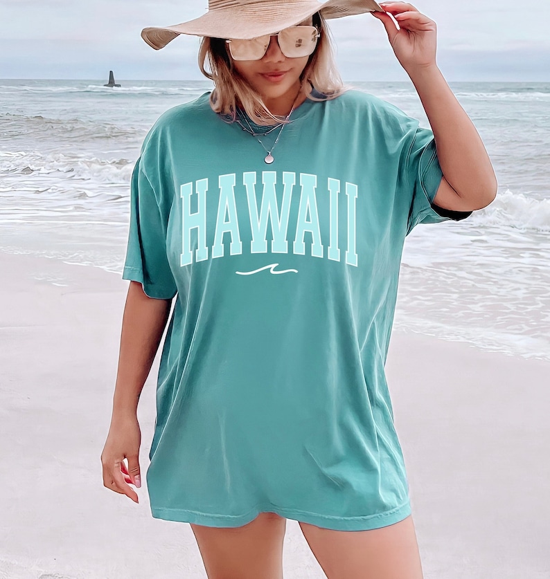 May include: A teal blue oversized t-shirt with the word "HAWAII" printed in a light blue color with a white wave underneath. The shirt is being worn by a person on a beach.