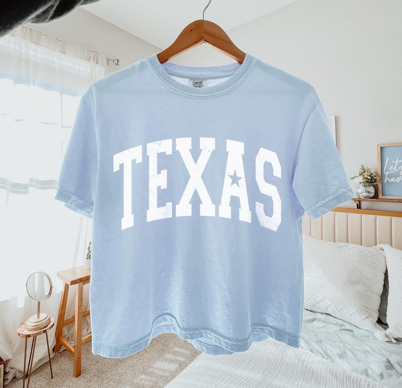 K&ouml;nnte beinhalten: Hellblaues, bauchfreies T-Shirt mit einem wei&szlig;en "TEXAS"-Aufdruck und einem kleinen Stern. Das Shirt hat einen Used-Look.