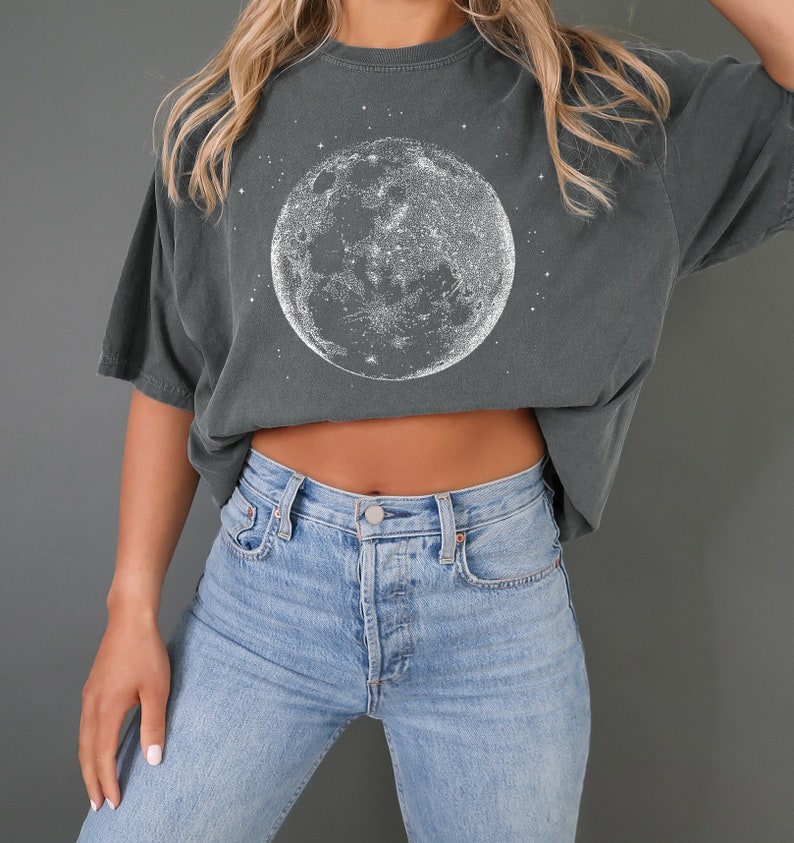 May include: Dark grey t-shirt with a white moon and star design. The t-shirt has a relaxed fit and short sleeves. The model is wearing light blue jeans.