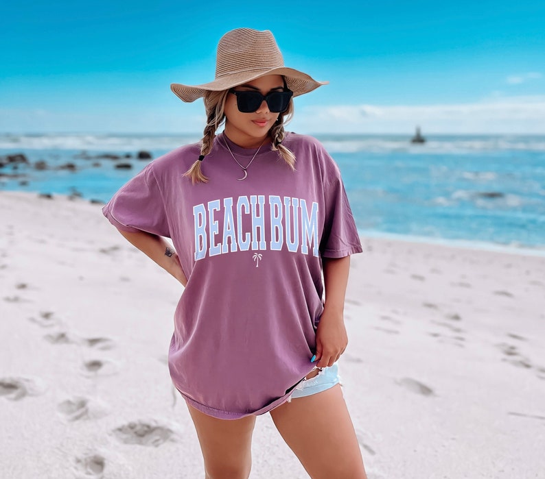 以下が含まれることがあります： 紫色のオーバーサイズのTシャツを着た女性。Tシャツには白と青の文字で「ビーチバム」と書かれている。彼女は麦わら帽子とサングラスをかけている。背景は青い海のあるビーチである。