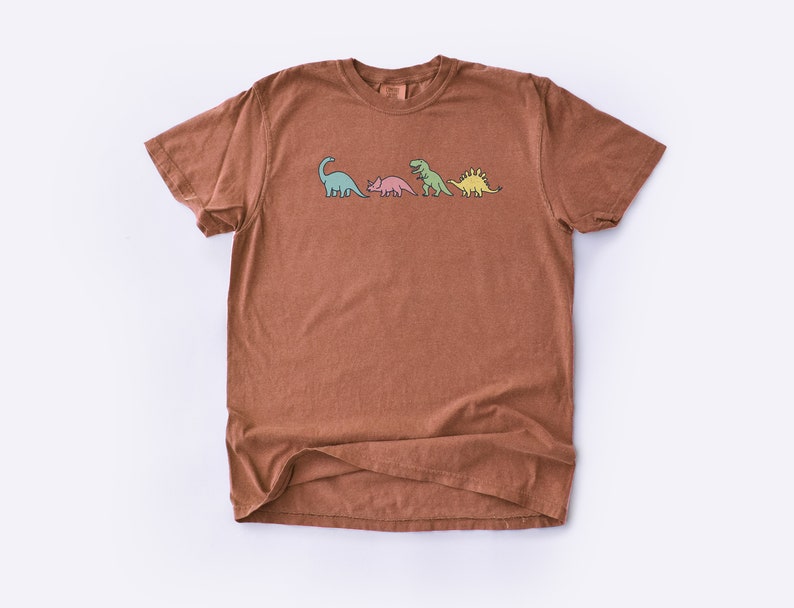 May include: A brown t-shirt with a graphic of four dinosaurs in a line. The dinosaurs are green, pink, green, and yellow.