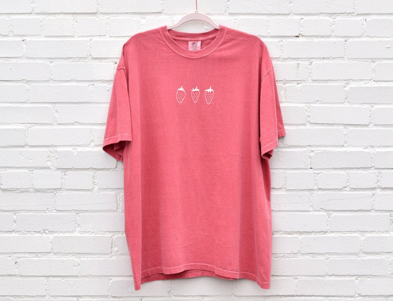 Op de afbeelding: Een koraalroze T-shirt met een wit geborduurd ontwerp van drie aardbeien.