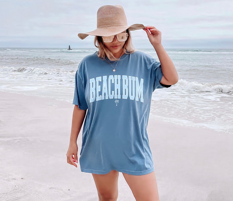 以下が含まれることがあります： ビーチで、白い文字で「ビーチバム」と書かれた青いTシャツを着た女性。彼女は麦わら帽子とサングラスをかけています。