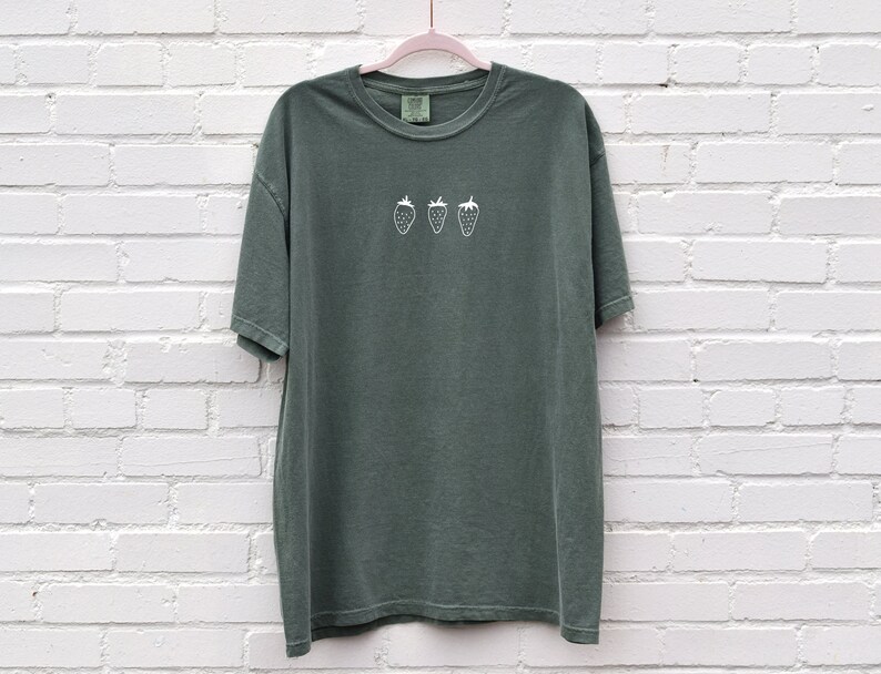Op de afbeelding: Een donkergroen T-shirt met een wit geborduurd ontwerp van vier aardbeien.
