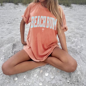 以下が含まれることがあります： ビーチで座っている女性。彼女はピーチ色のTシャツを着ています。Tシャツには青い文字で「BEACHBUM」と書かれており、くたびれた感じとヤシの木のグラフィックが施されています。